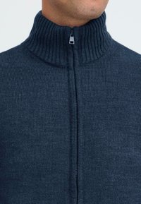 Nærbilde av en person som har på seg en mørk blå ribbestrikket hettegenser med høy krage, som er lukket opp til halsen.