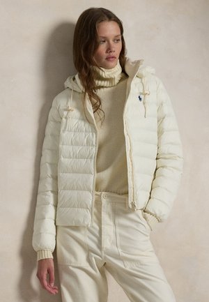 Γυναίκα που φοράει κρεμ καπιτονέ μπουφάν πάνω από ζιβάγκο και ταιριαστά ψηλόμεσα παντελόνια, στέκεται μπροστά σε έναν απλό μπεζ τοίχο.