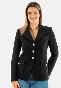 Blazer negro con solapa con muesca, cinco botones plateados y dos bolsillos frontales. Tela suave con un ajuste entallado. Se lleva sobre una blusa clara.