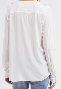 Blouse blanche à manches longues avec une texture lisse, présentant un empiècement dans le dos et des accents de boutons sur les manches. Ourlet arrondi.