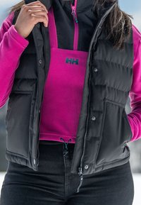 Veste matelassée noire sans manches portée sur un haut en polaire rose vif, avec une fermeture éclair à l'avant et des poches latérales, sur un fond hivernal.