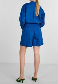 Koboltinsininen paita takana laskoksella, yhdistetty samansävyisiin shortseihin. Vihreät korkokengät. Sileä kangas, rento istuvuus, lyhyet hihat.