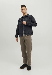 Melna džinsa jaka ar krūšu kabatām, kombinēta ar gaiši bēšām biksēm. Modelis stāv, demonstrējot brīvu piegriezumu un pogu aizdari. Melni apavi.