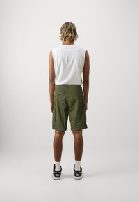 Person med kort dreadlock-hår som bär en ärmlös vit skjorta, gröna cargoshorts, vita strumpor och svarta sneakers, stående med ryggen vänd.
