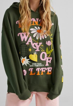 Sweat à capuche vert avec des fleurs colorées, des cœurs et un texte disant "Think way of life. Feed your mind positivity." Personne portant un pantalon rose.