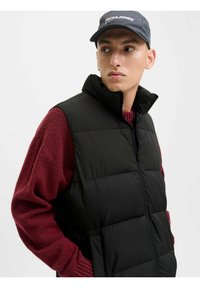 Chaleco acolchado negro con alto cuello, cierre de cremallera, combinado con un suéter rojo texturizado; el modelo lleva una gorra azul marino con logo.