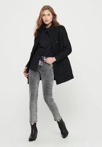 Manteau noir texturé avec des double boutons, superposé sur une chemise noire. Assorti à un jean droit gris délavé et des bottes à talons noires.