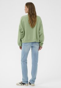 Femme aux longs cheveux bruns portant un sweat-shirt vert clair, un jean bleu clair et des baskets blanches, se tenant de dos sur un fond blanc.