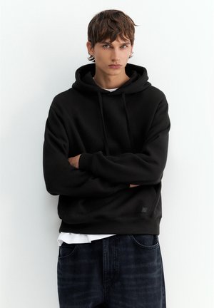 Jeune homme aux cheveux courts bruns portant un sweat à capuche noir et un jean foncé, debout les bras croisés sur un fond blanc.