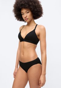 Γυναίκα με αφροκράνια, φοράει μαύρο ασύρματο σουτιέν και μαύρο σλιπ που ταιριάζει, στέκεται μπροστά σε λευκό φόντο.