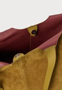 Schoudertas van leer en suède met bordeauxrode, gele en olijfgroene accenten; voorzien van een drukknoopsluiting en textuurrijke oppervlakken.