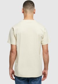 Mann med kort hår iført en enkel beige T-skjorte og blå jeans, vist fra ryggen mot en lys grå bakgrunn.