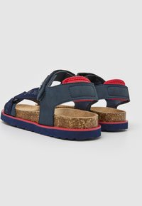 Mėlynos ir raudonos šlepetės su kamštiniu padu, guminėmis apačioje ir reguliuojamomis dirželiais. Yra tekstūruotas audinys ir putų akcentai.