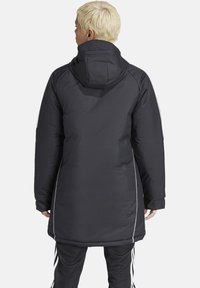 Schwarze gepolsterte Jacke mit Kapuze, versehen mit weißen Seitenstreifen, in mittellanger Ausführung und elastischen Bündchen. Glatter, strukturierter Stoff.