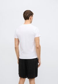 Λευκό βαμβακερό T-shirt με κοντά μανίκια, στρογγυλή λαιμόκοψη και λεία υφή, συνδυασμένο με σκούρο σορτς που έχει ελαφριά γυαλάδα. Πλάγια όψη από πίσω.