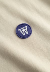 Beige stof med et rundt lilla broderet mærke med et hvidt stiliseret "W" symbol. Glat tekstur, minimalistisk design.