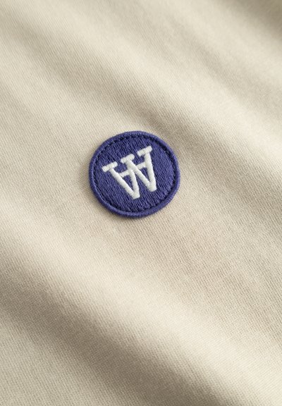 Tissu beige avec un patch rond brodé violet arborant un symbole "W" stylisé en blanc. Texture lisse, design minimaliste.
