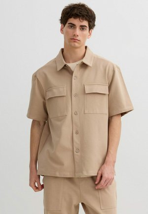 Joven con camisa beige de manga corta abotonada con dos bolsillos en el pecho y pantalones beige a juego, posando frente a un fondo liso.