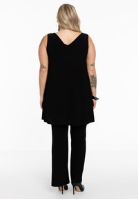 Schwarzes ärmelloses Tunika-Oberteil mit V-Ausschnitt auf der Rückseite, kombiniert mit schwarzen Hosen. Der Stoff wirkt glatt und hat einen lockeren Sitz sowie seitliche Schlitze. Schwarze High Heels.