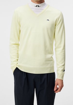 Mężczyzna w bladożółtym swetrze z dekoltem w serek nałożonym na biały golf z małymi czarnymi logo, w zestawieniu z ciemnymi plisowanymi spodniami.