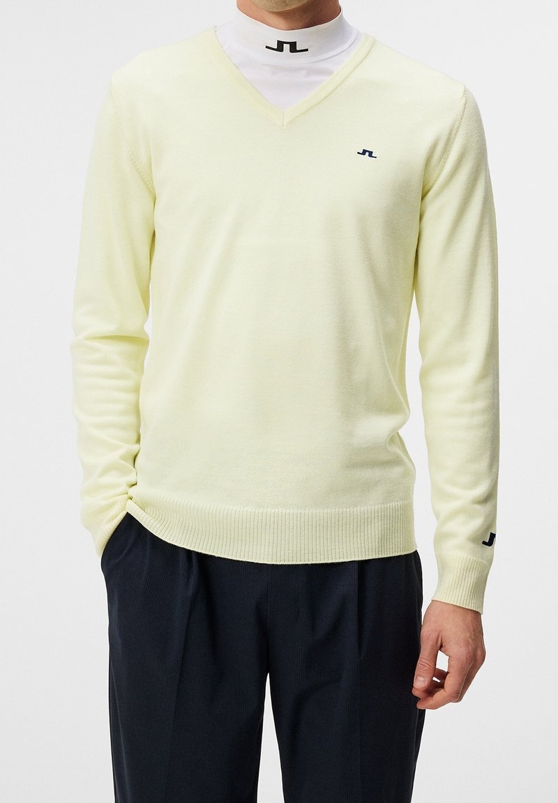 Homme portant un pull jaune pâle à col en V par-dessus un sous-pull blanc avec petits logos noirs, associé à un pantalon plissé foncé.