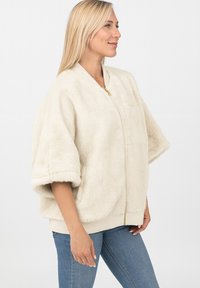 Veste bomber en fausse fourrure crème avec fermeture éclair, manches larges et poignets côtelés. Elle présente une texture douce et une coupe décontractée.