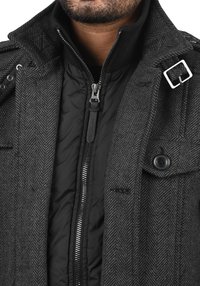 Grijze visgraatmantel met zwart doorgestikt interieur, voorzien van een ritssluiting, knoopdetail en een metalen accent op de kraag.