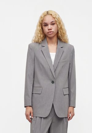 Jonge vrouw met krullend blond haar die een oversized lichtgrijze colbertjas en bijpassende broek draagt over een wit topje.