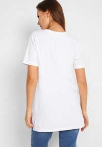 T-shirt bianca in cotone con scollatura rotonda, maniche corte e orlo posteriore più lungo; design semplice senza fantasie o accenti.