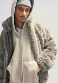 Jachetă din blană sintetică bej cu glugă, având fermoar în față și buzunare laterale, purtată peste un hanorac bej deschis. Textură moale și croială relaxată.