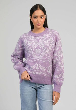 Femme portant un pull lavande avec des motifs floraux blancs et un jean bleu clair, debout devant un fond gris uni.