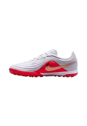 Chaussure de football basse blanche et rouge avec le logo Nike doré et une semelle texturée pour gazon, vue de côté.