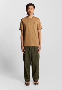 Brun bomullströja med korta ärmar och en liten gul logotyp på bröstet, i kombination med gröna cargobyxor och mönstrade bruna skor.