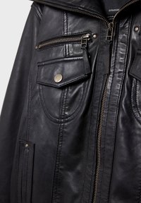 Veste en cuir noire en gros plan montrant la fermeture éclair, la poche poitrine zippée, la poche à rabat avec bouton et les détails cousus.