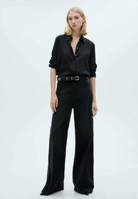 Camicia nera con bottoni e maniche arrotolate, abbinata a jeans in denim neri a gamba larga e alta vita. Include una cintura con fibbia in argento.