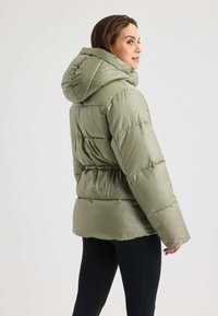 Giacca puffer khaki con cappuccio, motivo trapuntato, vita elasticizzata e superficie liscia; indossata con leggings neri.