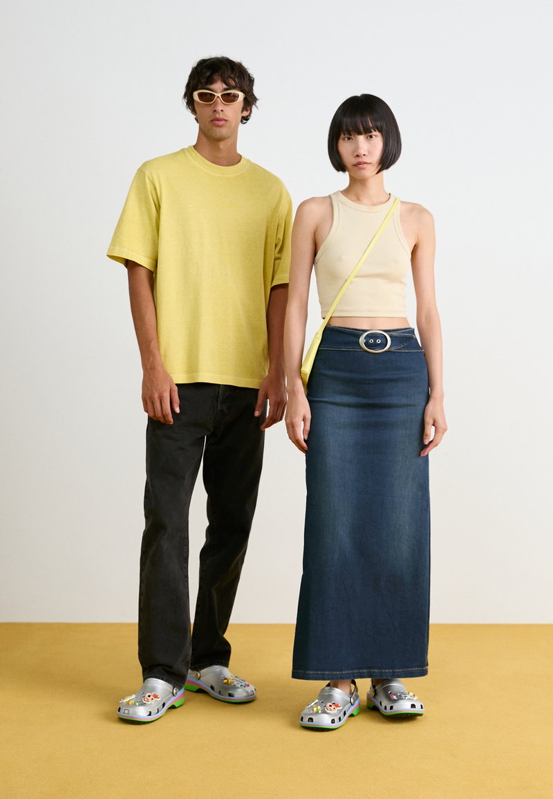 Gul oversized t-skjorte, beige tettsittende crop top, blå denim maxiskjørt, grå klogs med dekorative charms, på en nøytral bakgrunn.