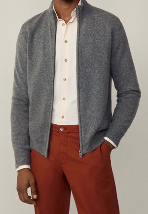 Mann trägt eine offene graue Strickjacke über einem weißen Hemd und rostfarbenen Hosen, eine Hand in der Tasche, vor schlichtem Hintergrund.