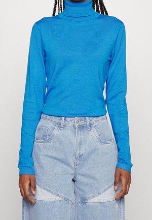 Kobieta ubrana w jasnoniebieski sweter z golfem i wysokie dwukolorowe jasnoniebieskie dżinsy, stojąca na jednolitym tle.