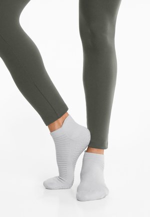 Ljusgrå ankelsockor med ribbat mönster och prickiga greppdetaljer, i kombination med åtsittande olivgröna leggings, som visar en stilren design.