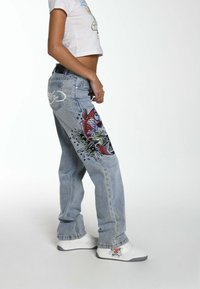 Model in a cropped white shirt and embellished jeans with fish designs, wearing white sneakers, poses against a plain background.
