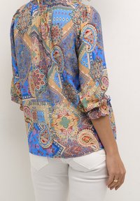 Blusa a fantasia colorata con tonalità di blu, beige e rosso. Ha maniche a tre quarti con polsini arricciati e una vestibilità comoda.