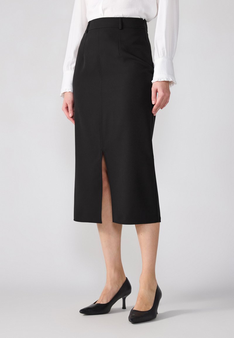 Jupe crayon noire mi-longue avec une fente avant, design ajusté, tissu lisse, associée à des chaussures à talons hauts noires. Blouse blanche visible.