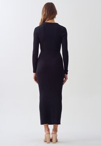 Kvinna som bär en långärmad, svart ribbad midiklänning och beige blockklackade sandaler, stående med ryggen mot kameran.