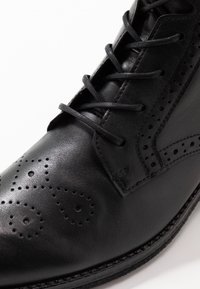 Svart lädersko med brogue-design som har dekorativa perforeringar, platta svarta snören och en slät yta.