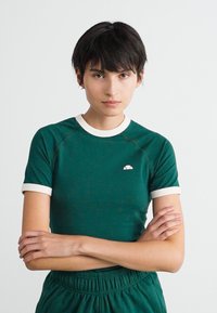 Kvinde med kort, mørkt hår iført en grøn t-shirt med hvid kant og matchende grønne bukser står med armene korslagte foran en ensfarvet baggrund.