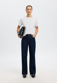 Wit T-shirt met korte mouwen gecombineerd met donkerblauwe, hooggetailleerde, wijd uitlopende jeans. Model houdt een zwarte tas vast met een uniek ontwerp. Zwarte schoenen.