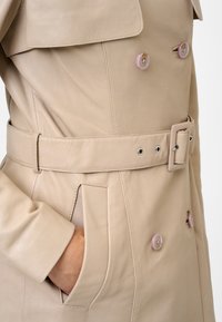 Beige leather trench coat featuring a double-breasted design, pink buttons, a belted waist, and side pockets with a smooth texture.