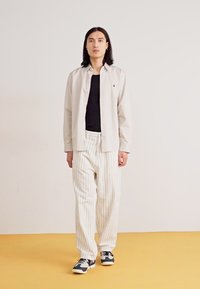 Helle beige Knopfleiste über einem schwarzen Shirt, kombiniert mit weißen Nadelstreifenhosen und grauen Sneakers. Weicher Stoff, lockere Passform.