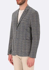 Blazer a quadri navy e beige con texture, chiusura a due bottoni, collo a rotondo e due tasche frontali. Abbinato a una camicia chiara.
